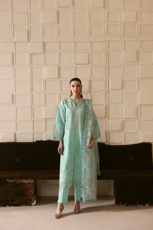 Woman in a light green traditional outfit standing against a textured wall.