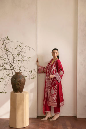 Woman in a red embroidered traditional outfit standing next to a vase with branches indoors.