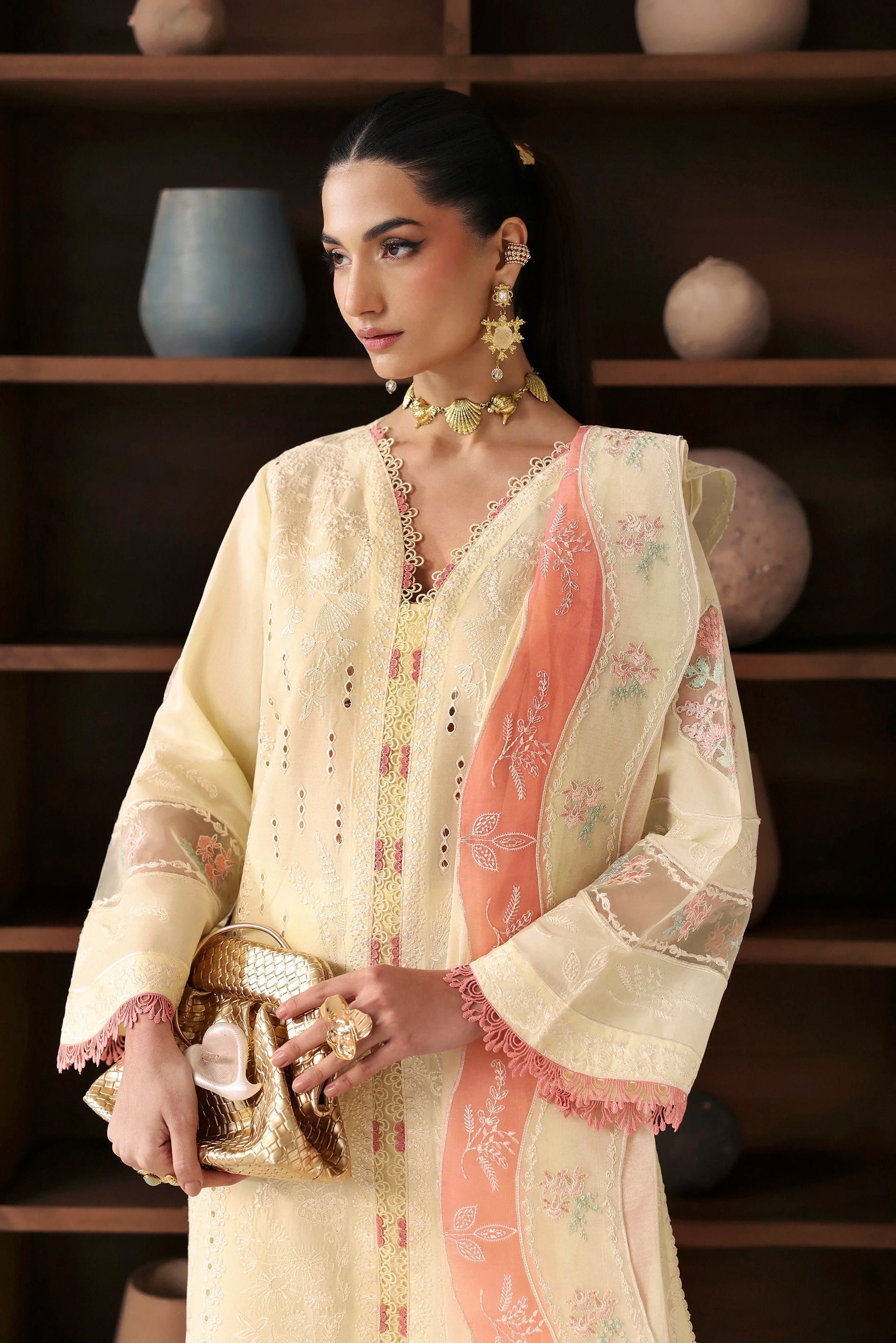 Woman in a traditional outfit with intricate designs, holding a decorative item, against a wooden shelf background.