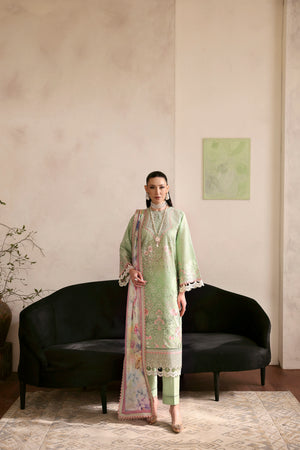 Woman in a light green traditional outfit with a floral dupatta standing in a room with a black sofa and a painting on the wall.