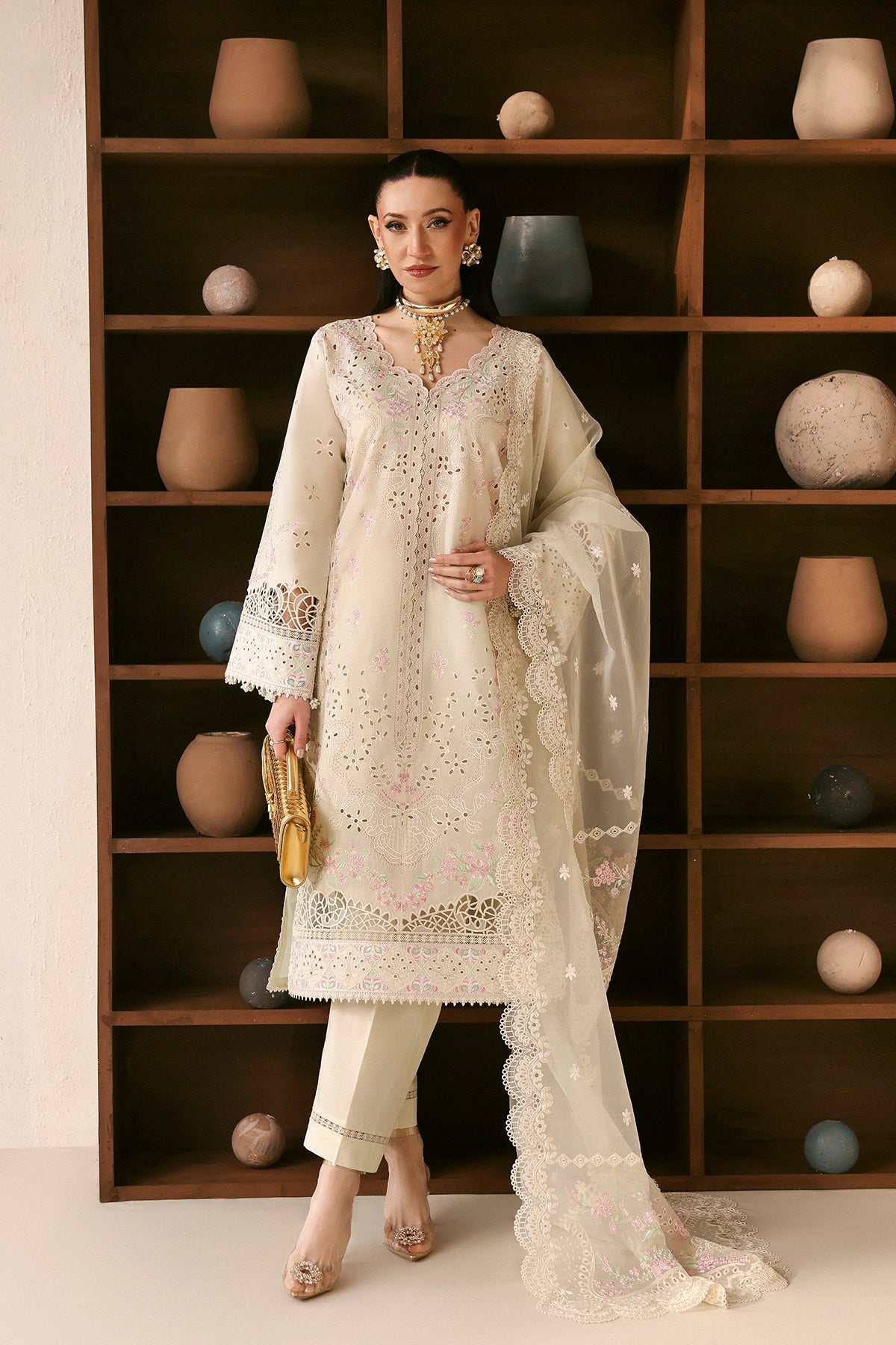 Woman in a cream embroidered outfit standing in front of a wooden shelf with decorative items.