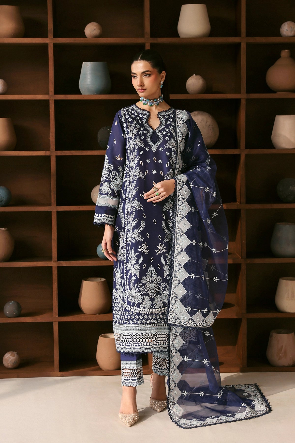 Woman wearing a blue embroidered traditional outfit in front of a wooden shelf with decorative pots.