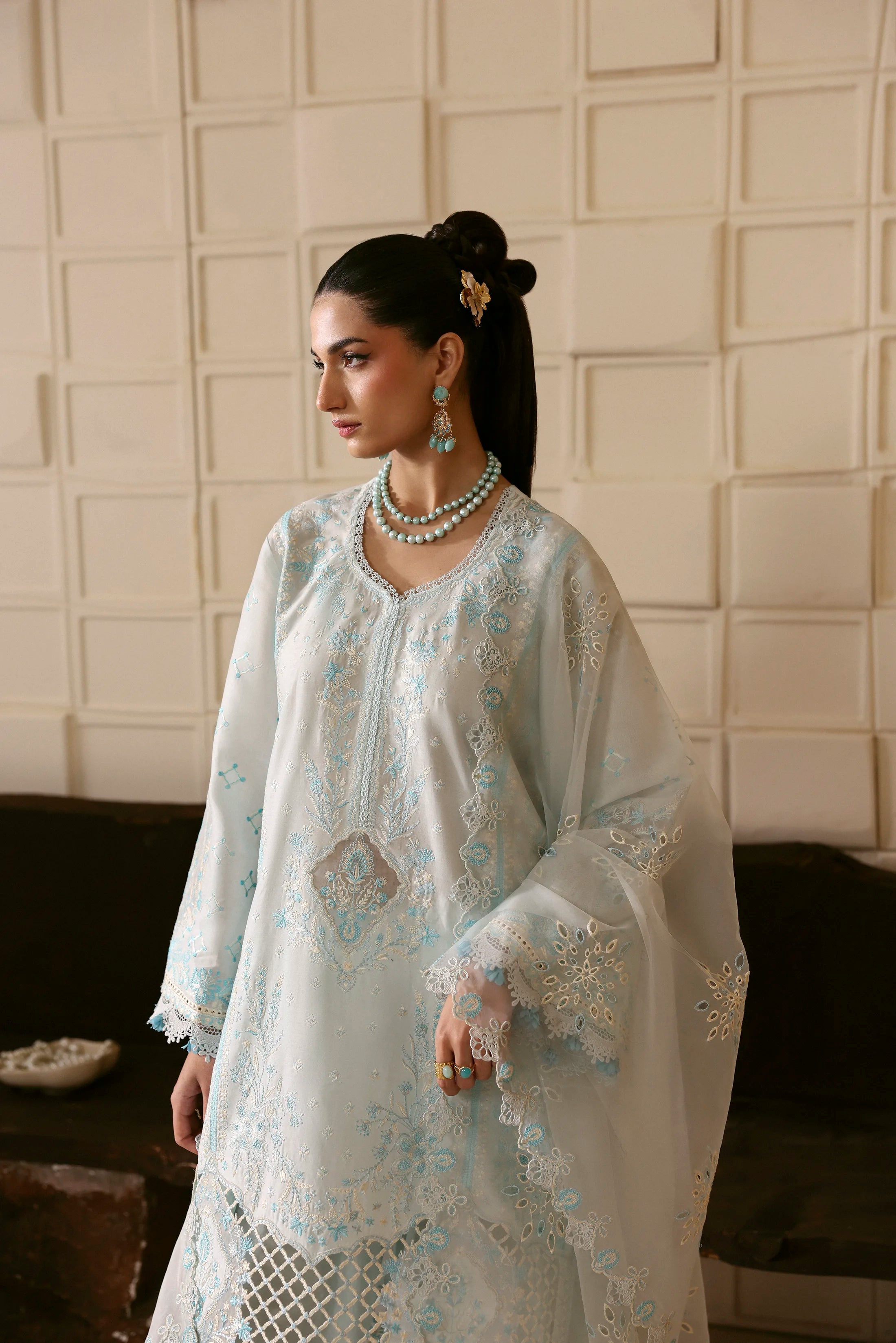 Woman wearing a light blue embroidered dress with jewelry against a neutral wall.