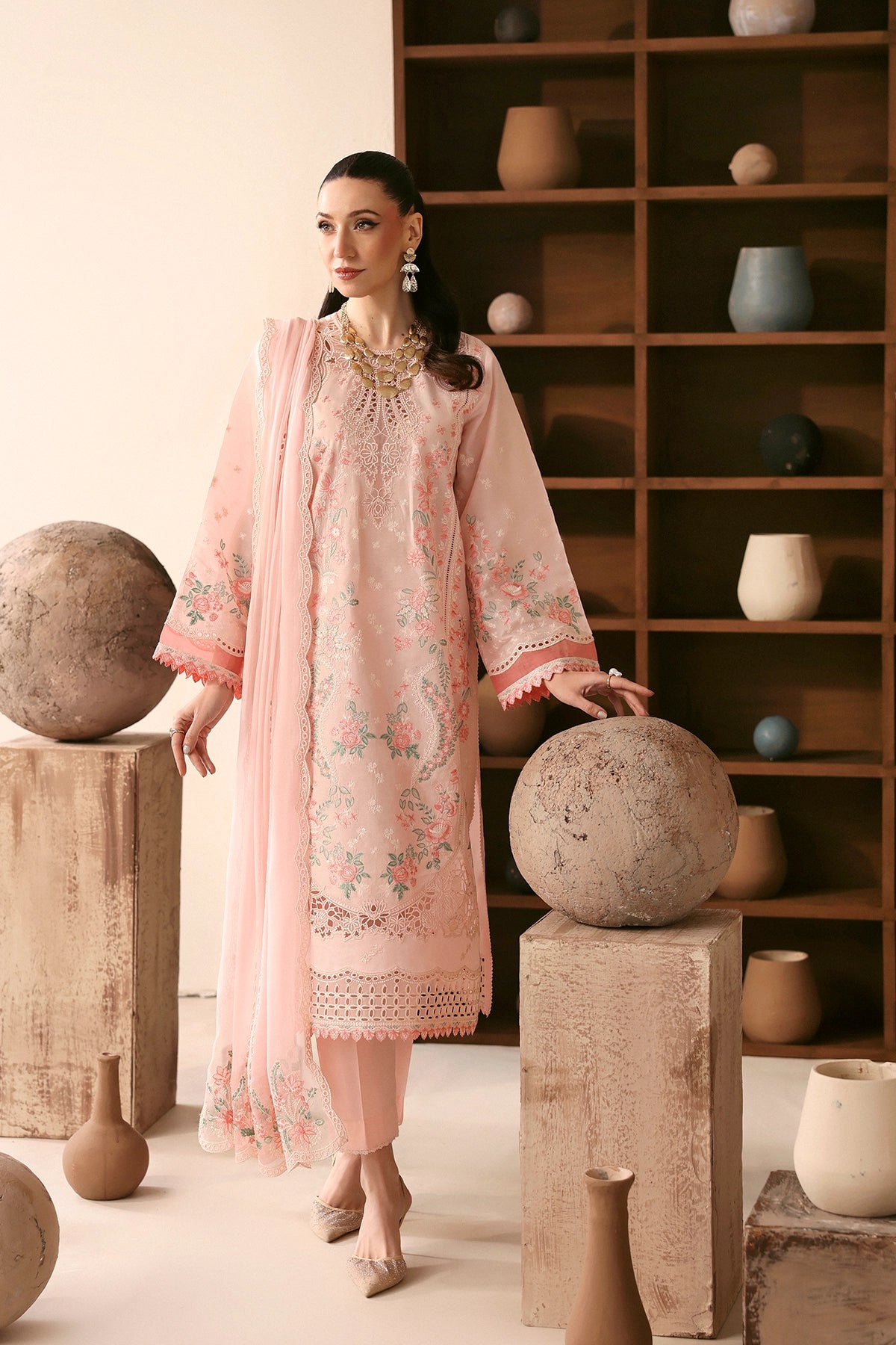 Woman in a pink embroidered outfit standing in a room with decorative shelves and spherical objects.