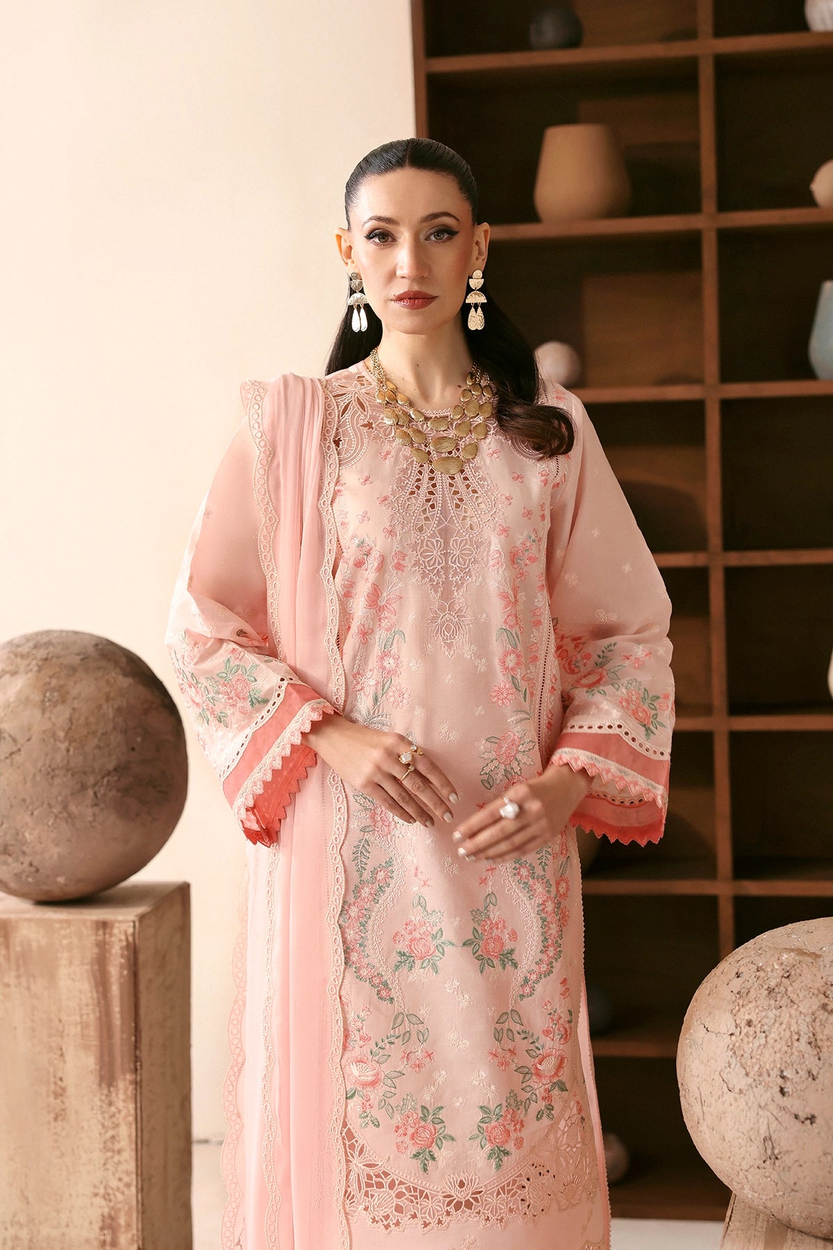 Woman in a light pink embroidered traditional outfit standing in a room with wooden shelves.