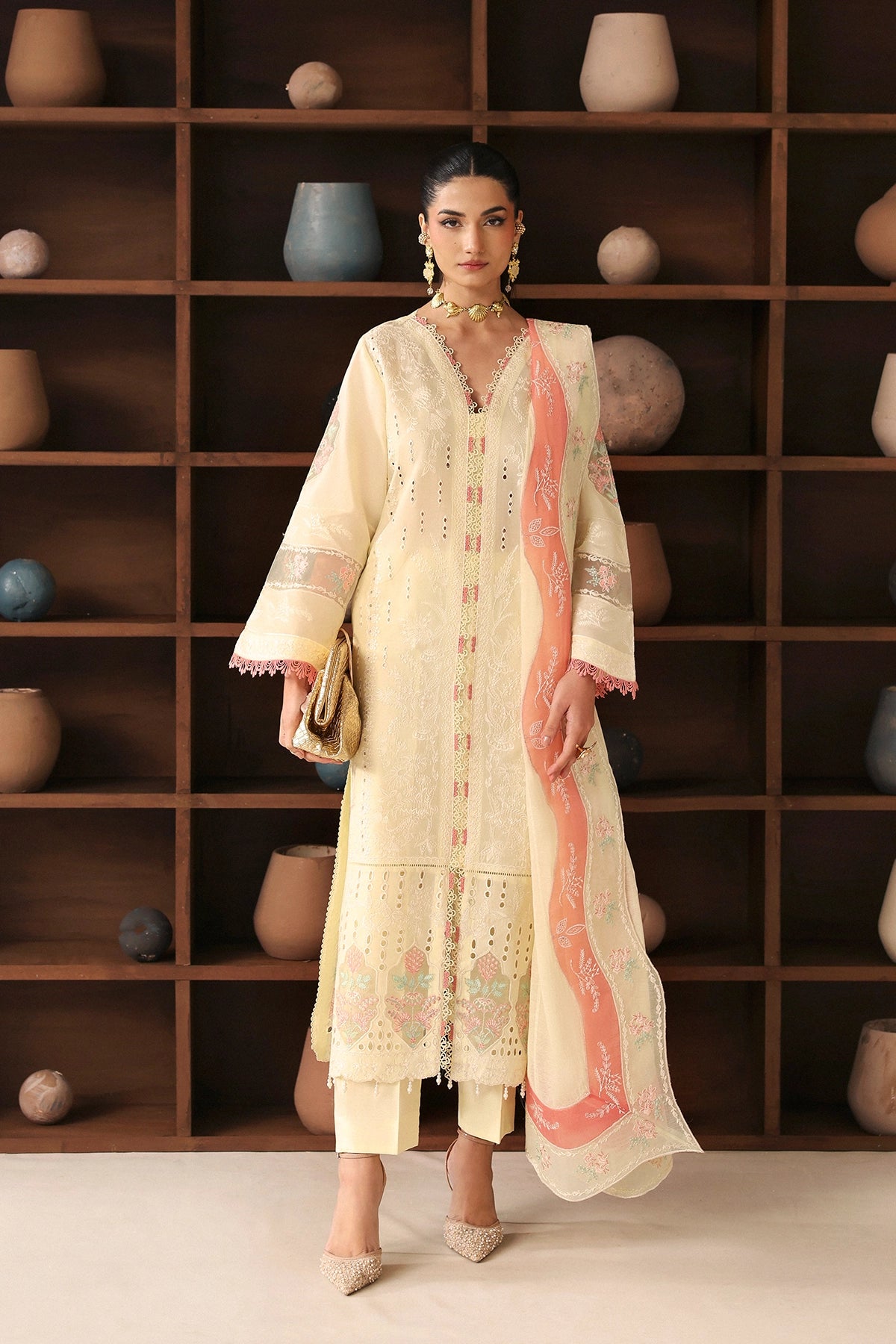 Woman in a cream embroidered outfit standing in front of a wooden shelf with decorative pots.