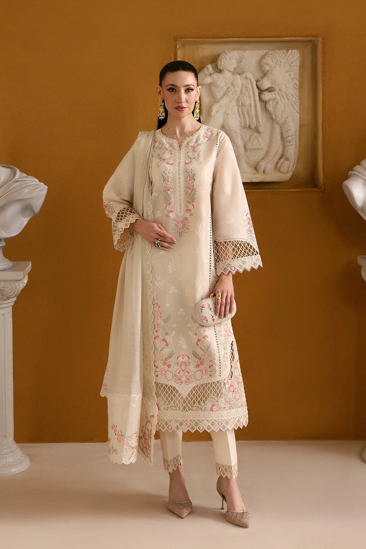 Woman in a cream embroidered outfit standing against an orange wall with decorative elements.
