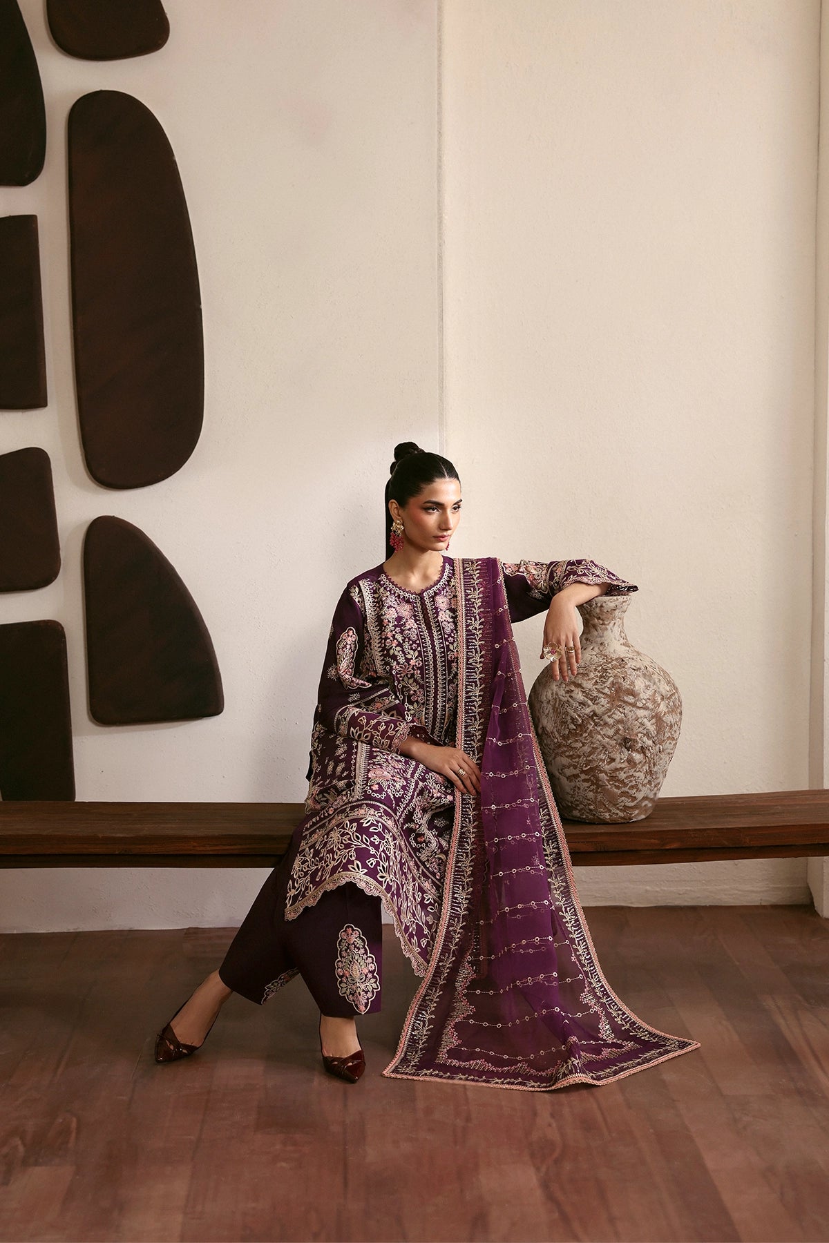 Woman in a purple and black traditional outfit sitting next to a decorative vase against a wall with abstract art.
