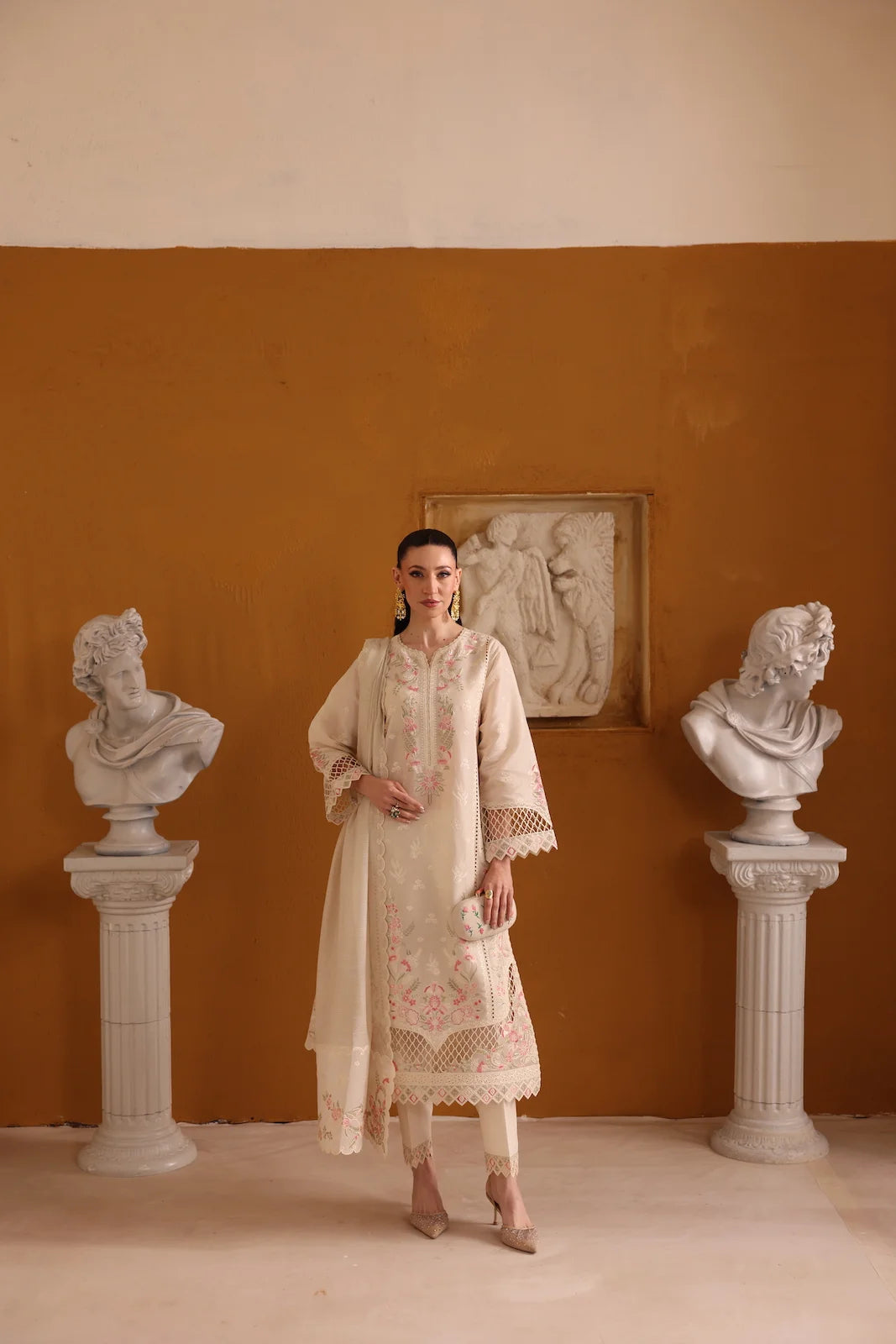 Woman in a cream embroidered outfit standing between classical busts against an orange wall.
