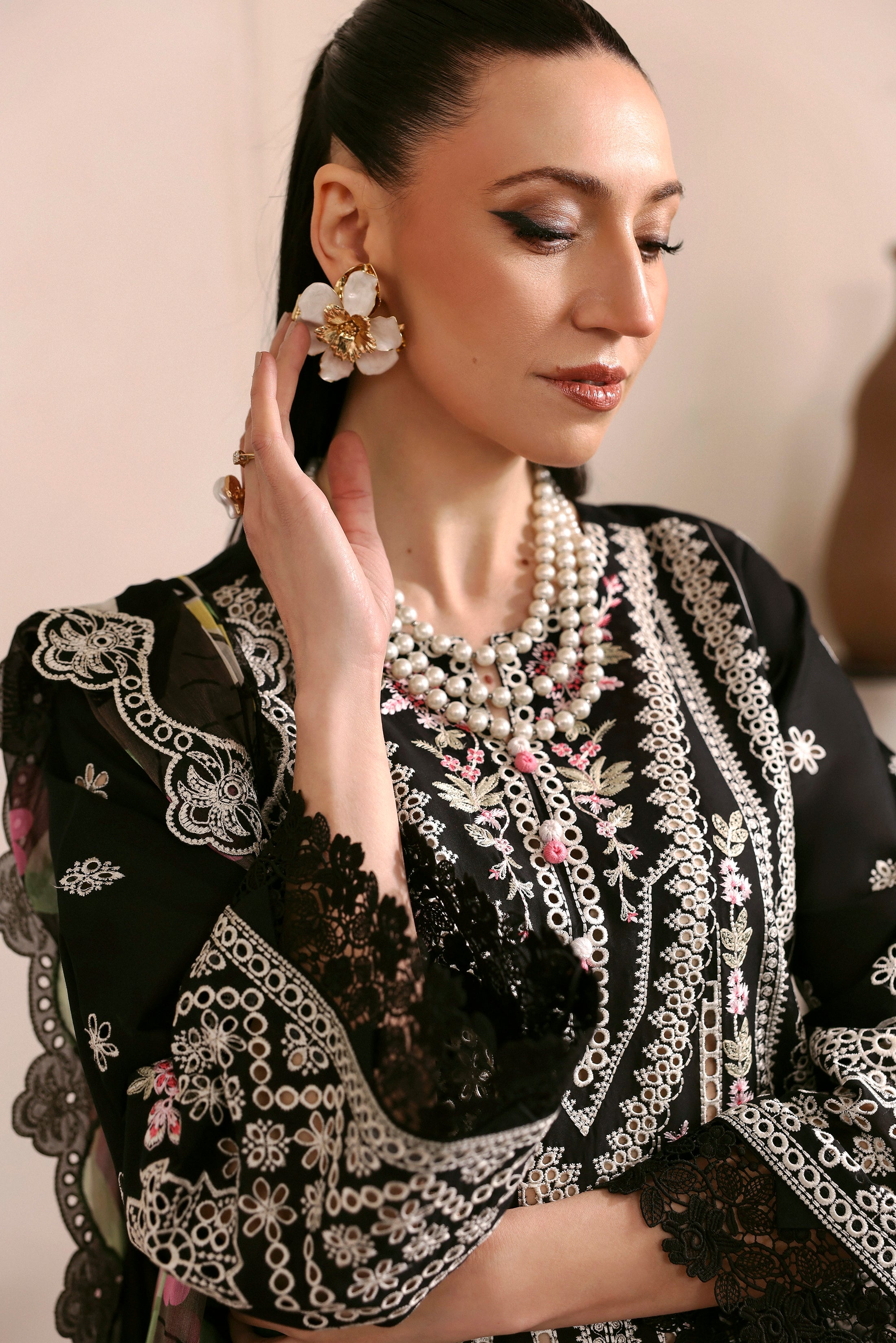 Woman wearing a black embroidered outfit with pearl jewelry against a neutral background