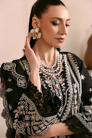 Woman wearing a black embroidered outfit with pearl jewelry against a neutral background