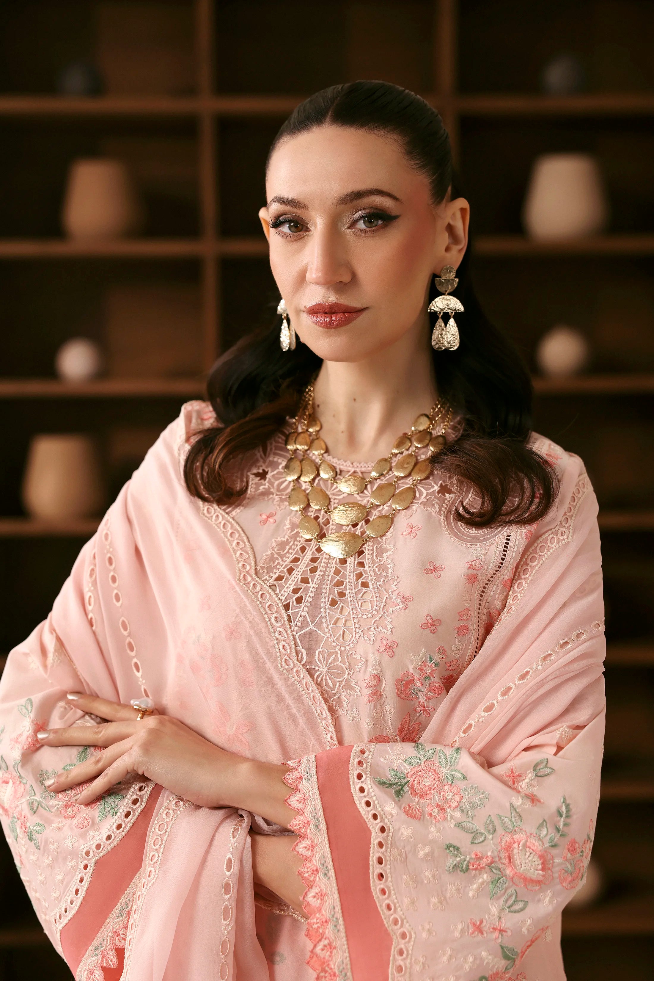 Woman wearing a pink embroidered outfit with gold jewelry against a wooden shelf background