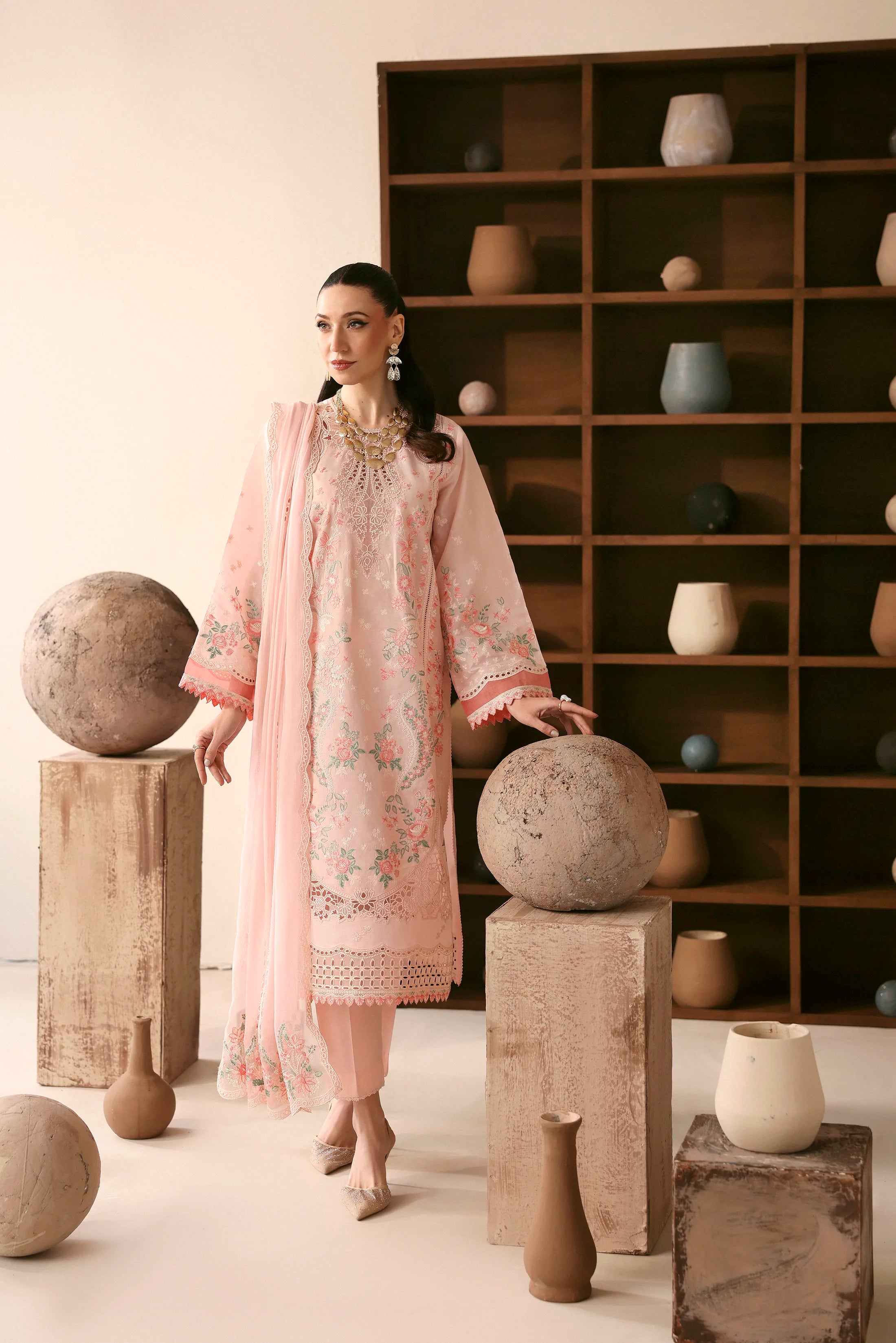 Woman in a light pink embroidered dress standing between stone spheres with a bookshelf in the background.
