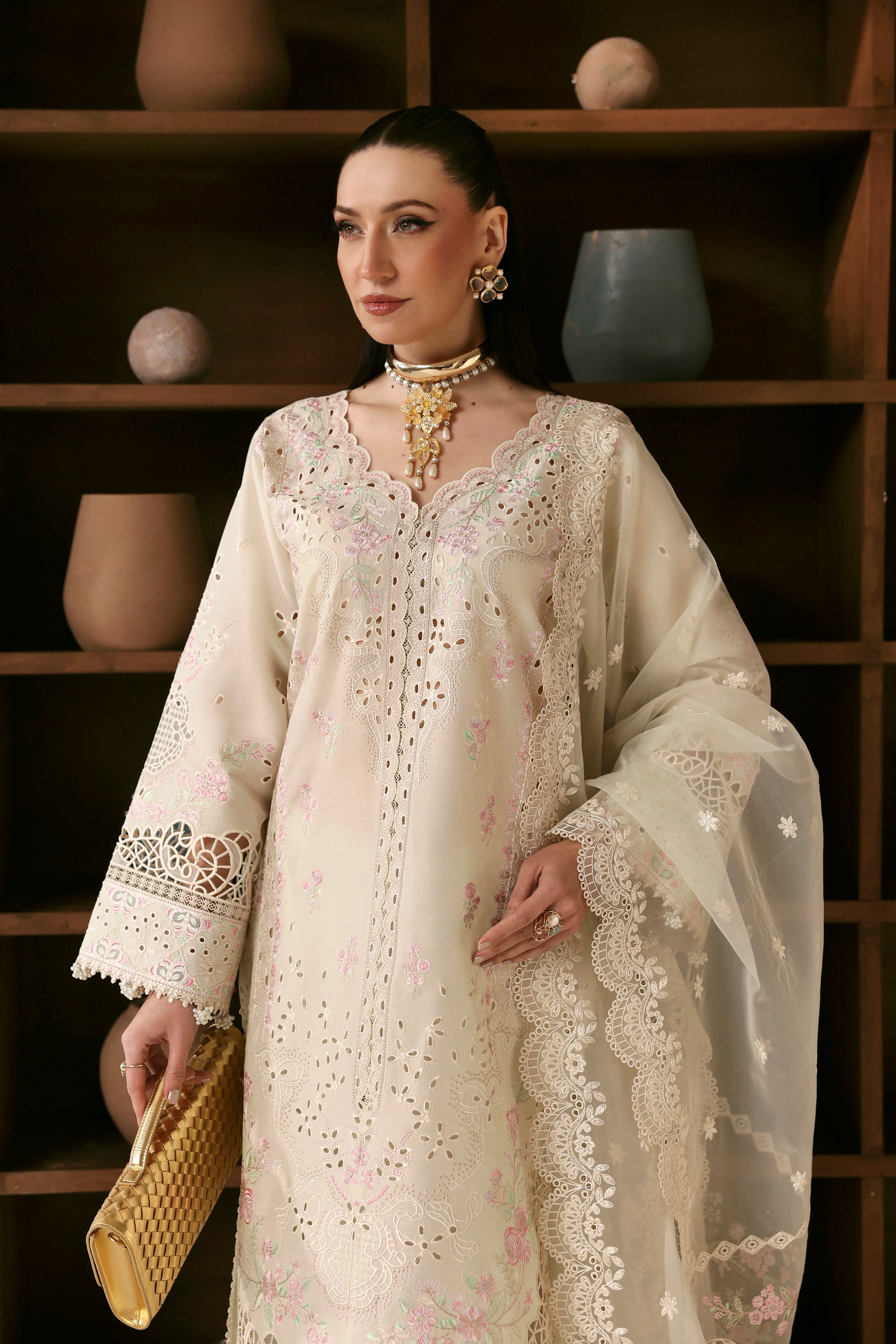 Woman wearing a cream embroidered dress against a wooden shelf background