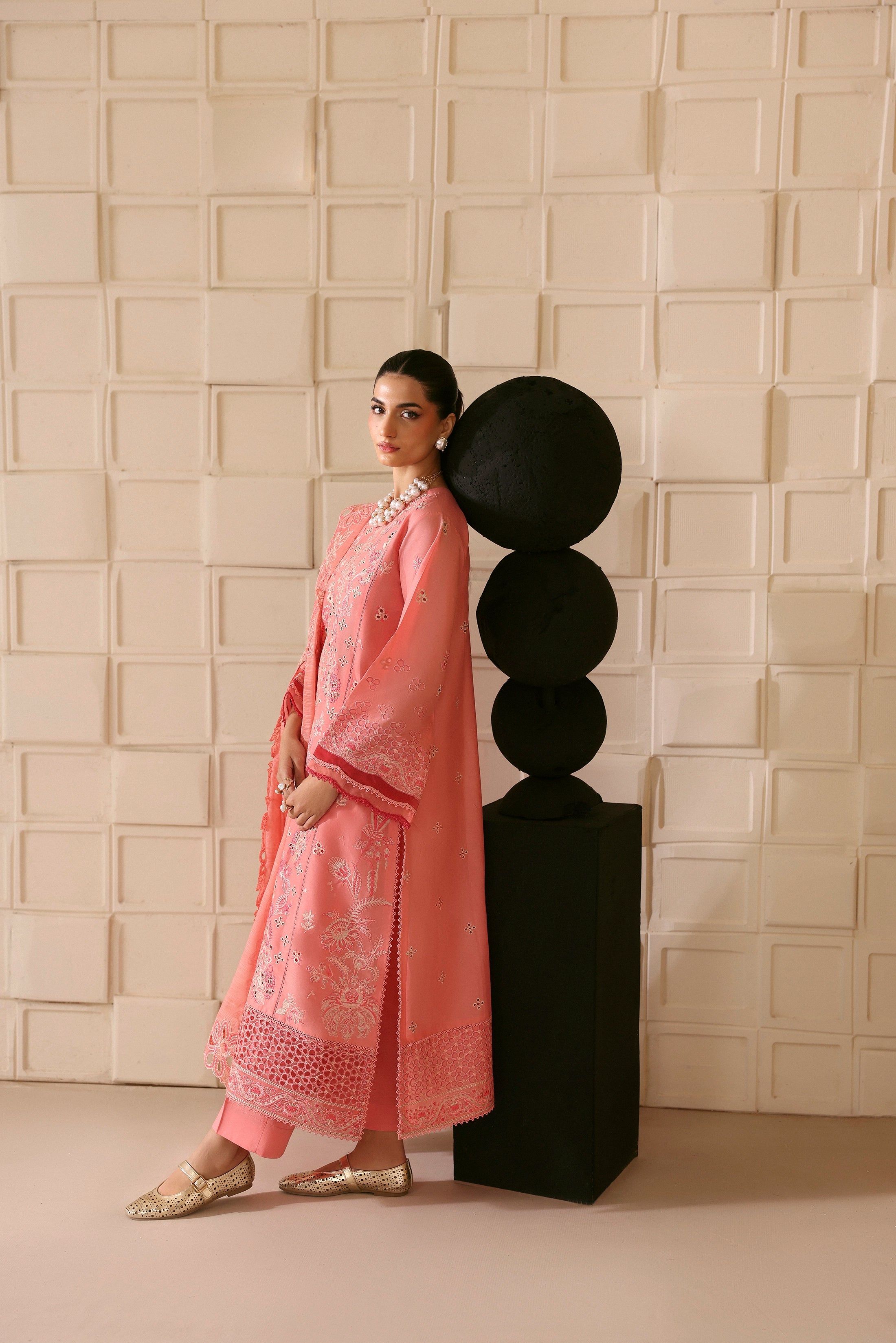 Woman in a pink traditional outfit standing next to a black decorative piece against a textured beige wall.