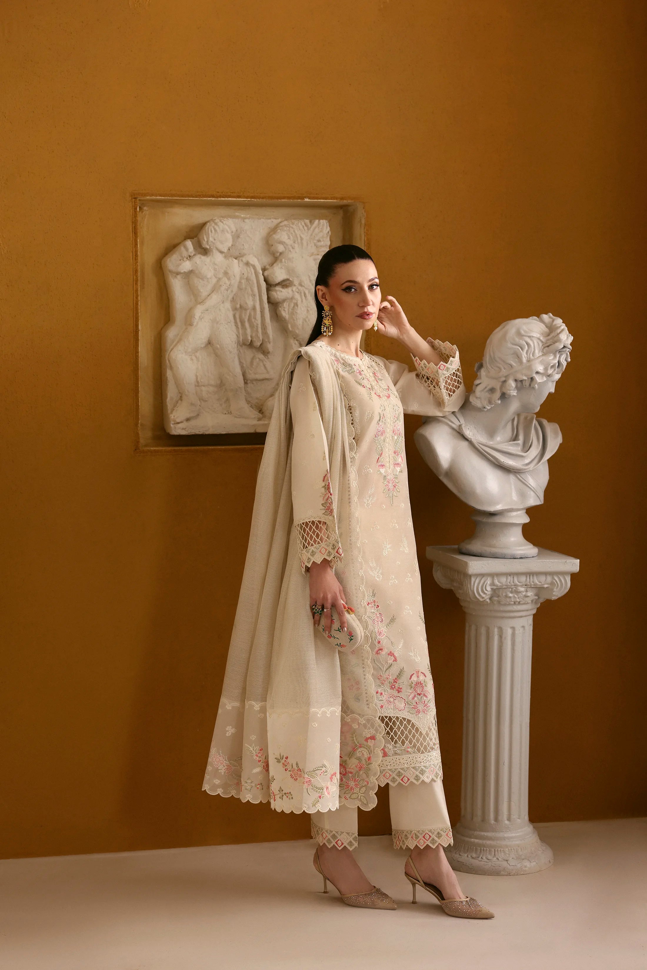 Woman in a cream embroidered outfit standing in a room with classical sculptures and wall art.