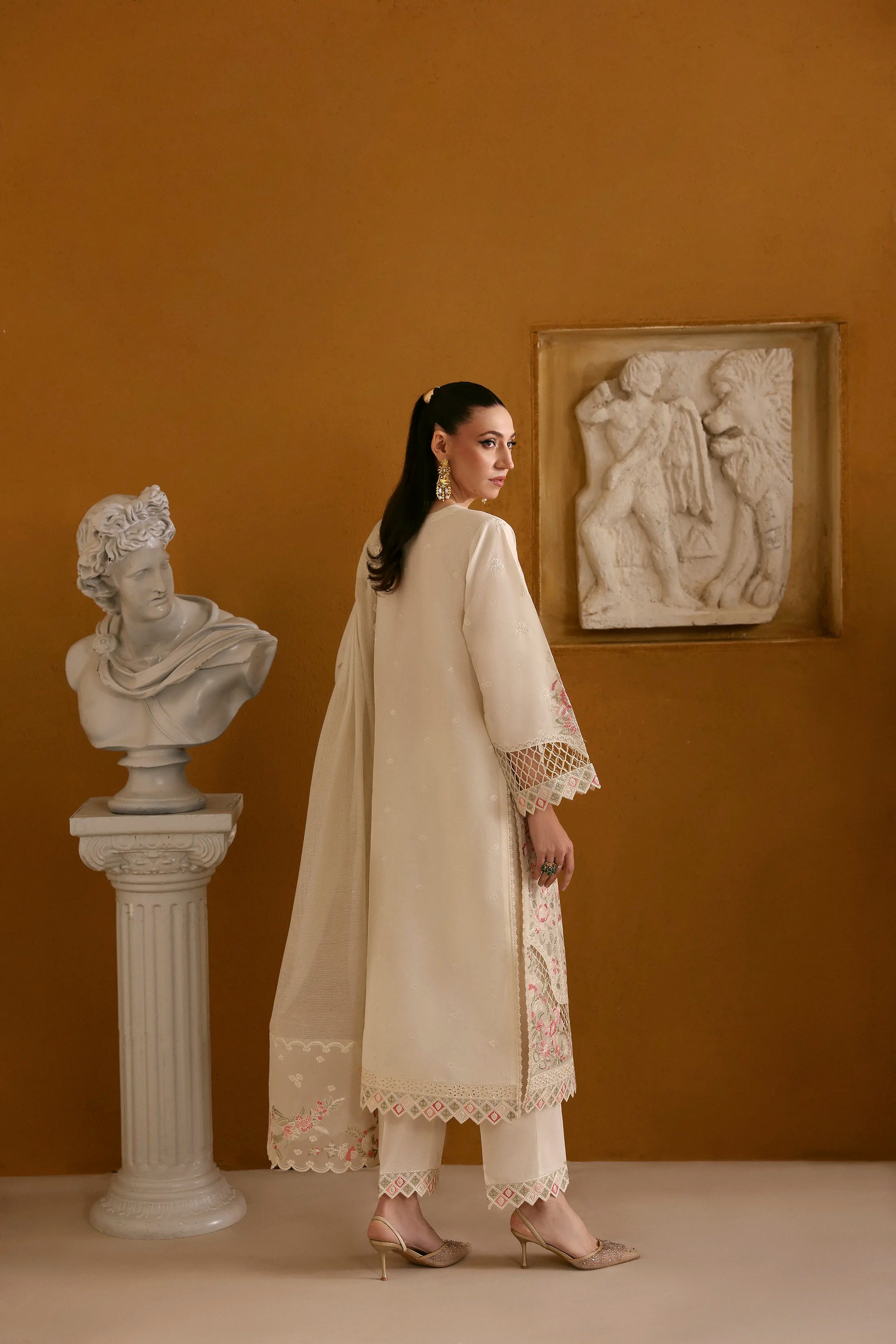 Woman in a cream traditional outfit standing in an elegant room with classical sculptures.