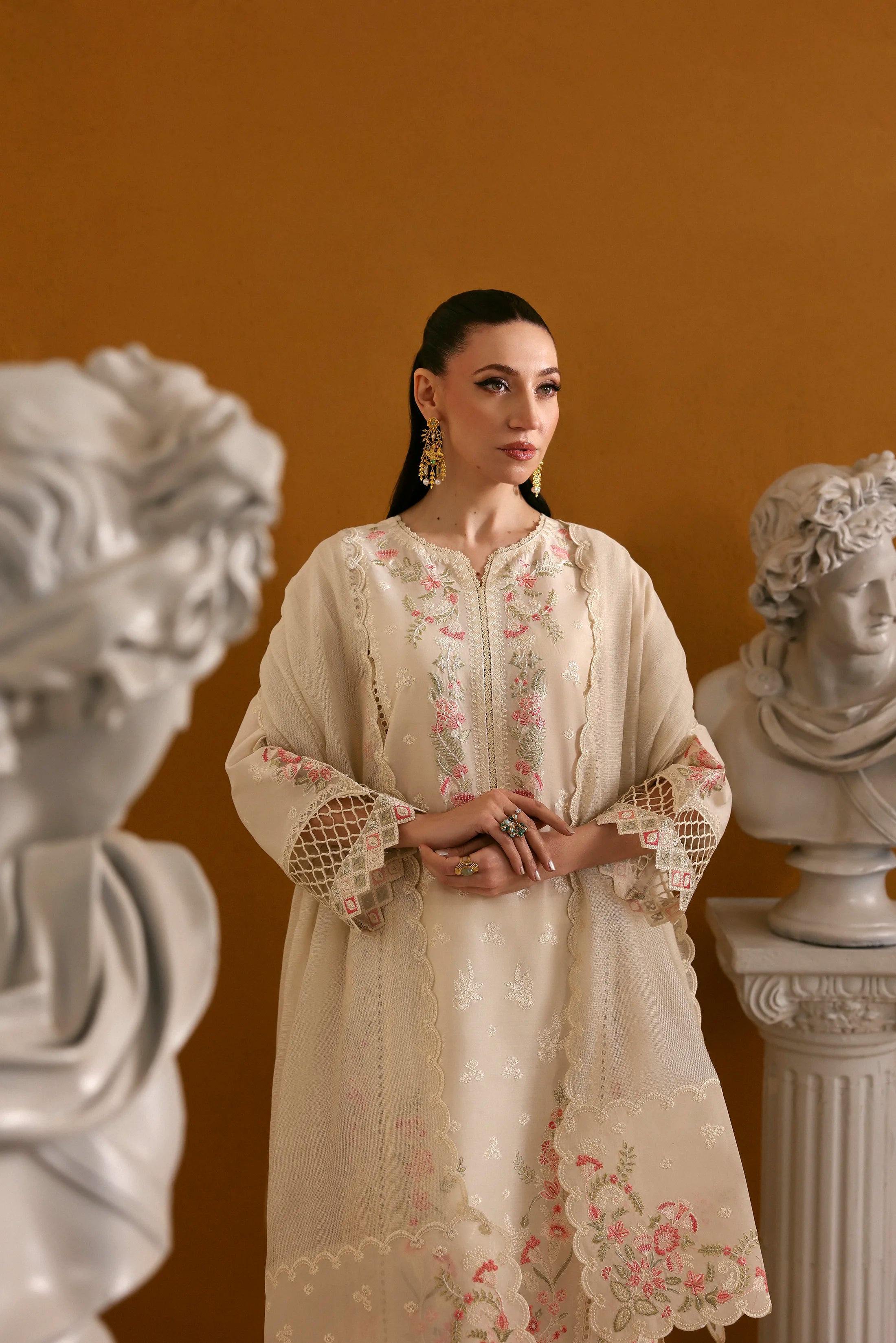 Woman in an embroidered dress standing between classical busts against a brown background