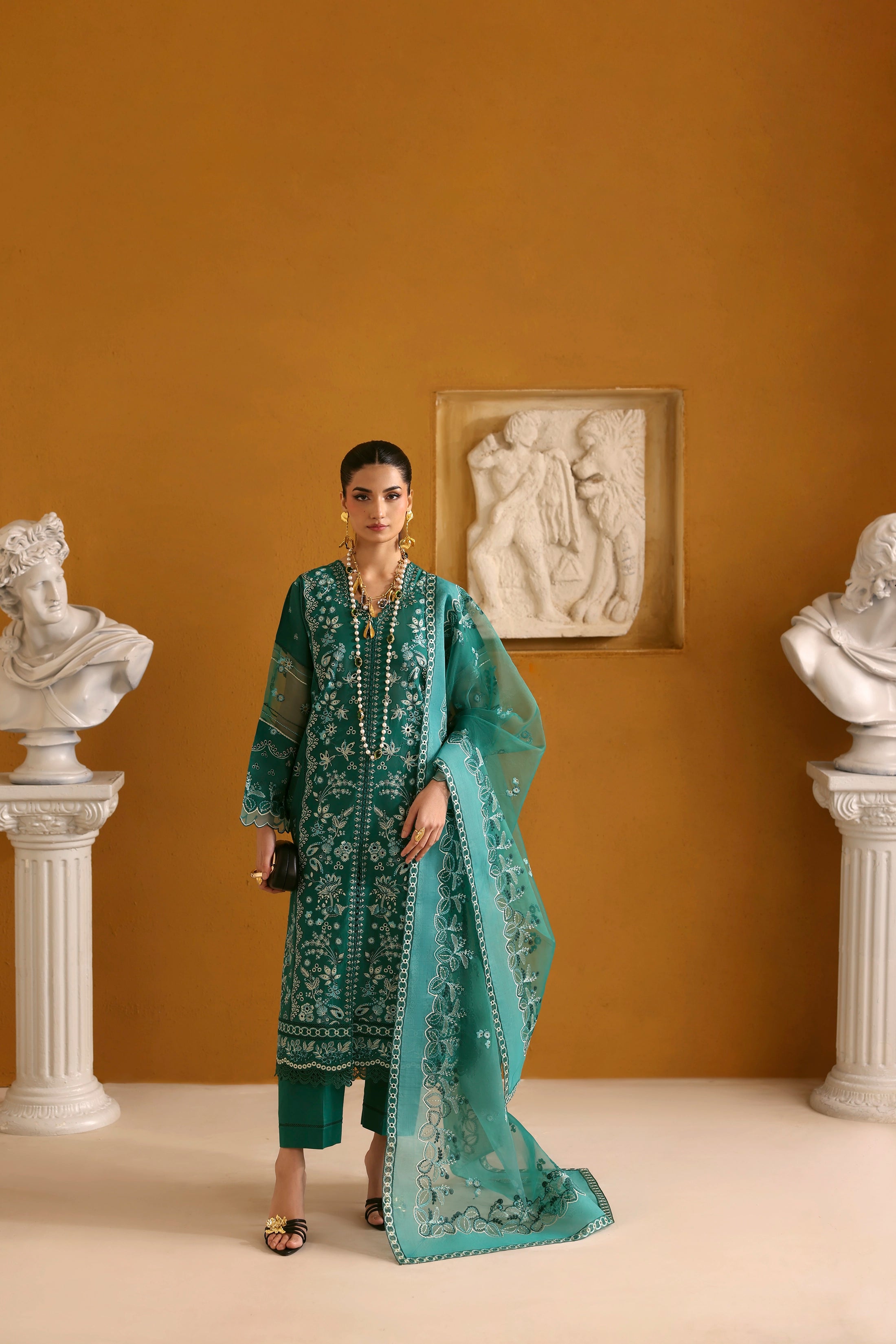 Woman in a teal embroidered outfit standing in a room with classical sculptures and wall art.