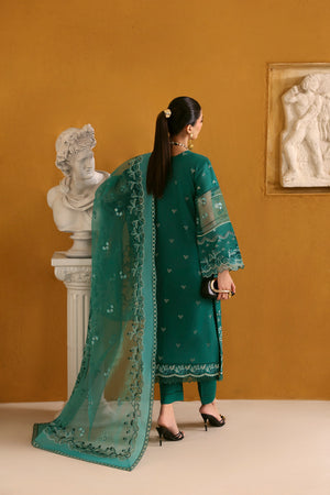 Woman in a teal traditional outfit with a matching dupatta, standing against a beige wall with sculptures.