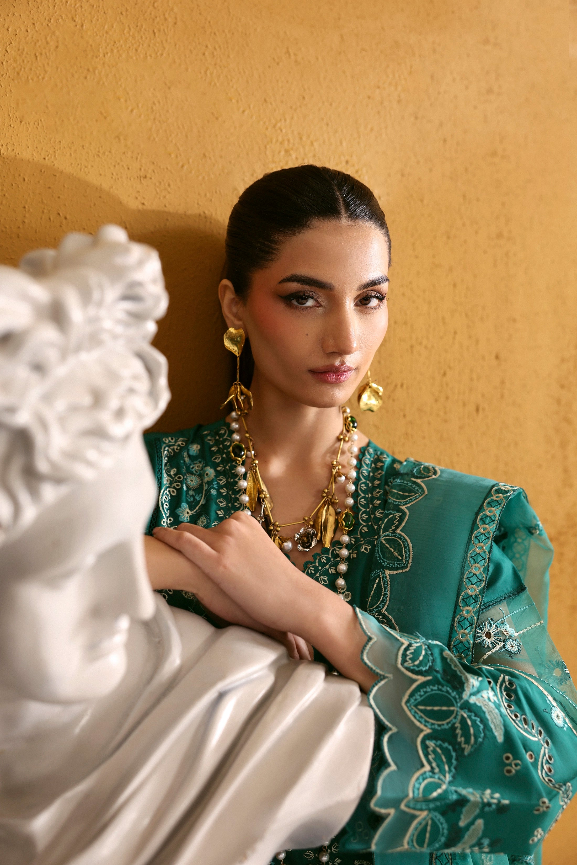 Woman in a teal embroidered outfit standing next to a classical statue against a beige wall.