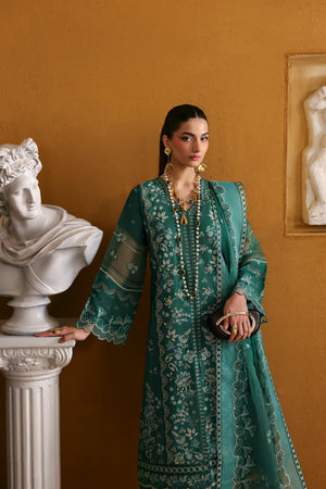 Woman in a teal embroidered dress standing against a beige wall with classical sculptures.